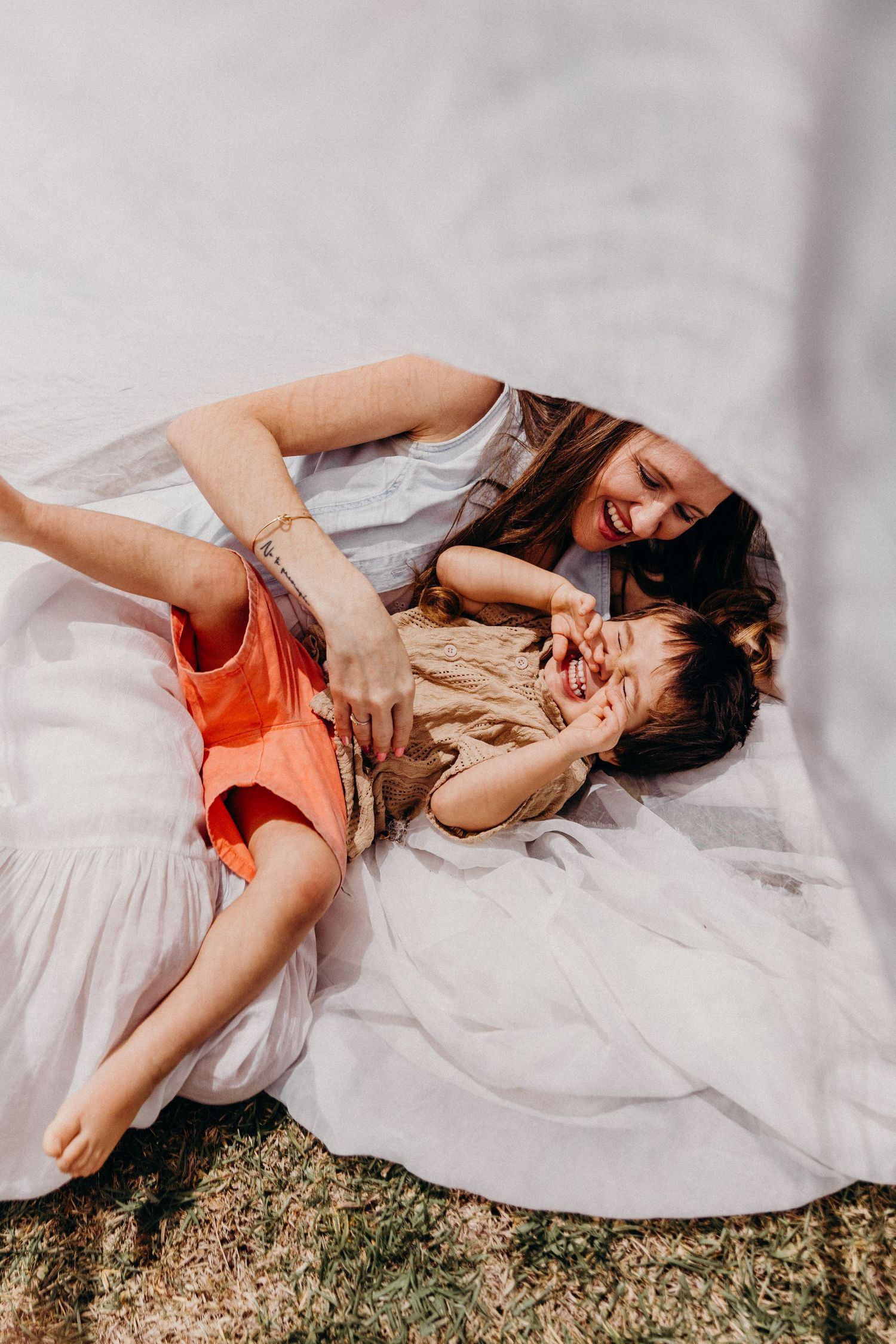 A mother and child laughing together while lying on a white blanket outdoors in warm sunlight.