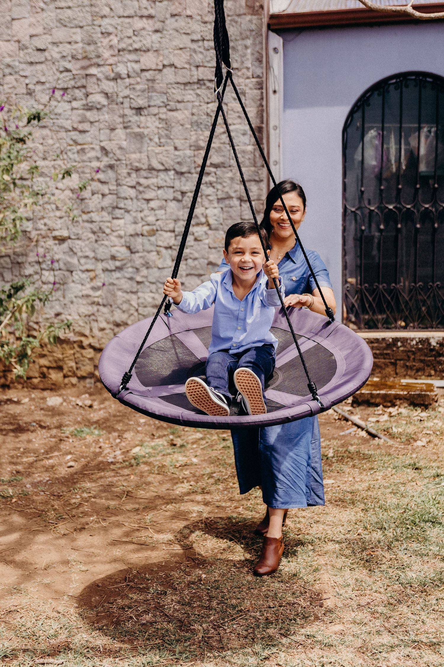 A smiling boy sits on a round nest swing while a woman pushes him in a sunny backyard.