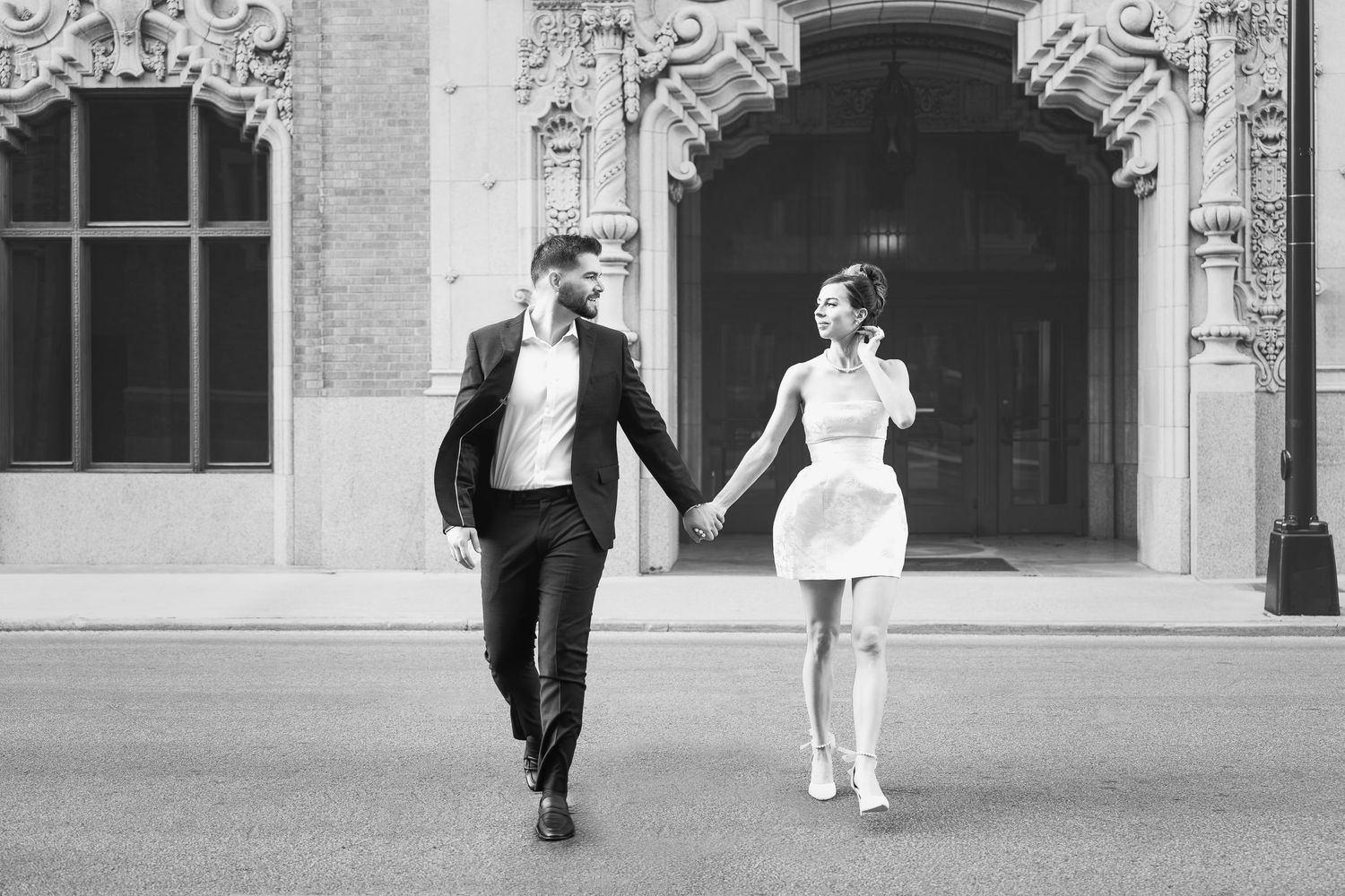 man in black suit holding fiancé's hand as they look at each other and cross the street in downtown San Antonio