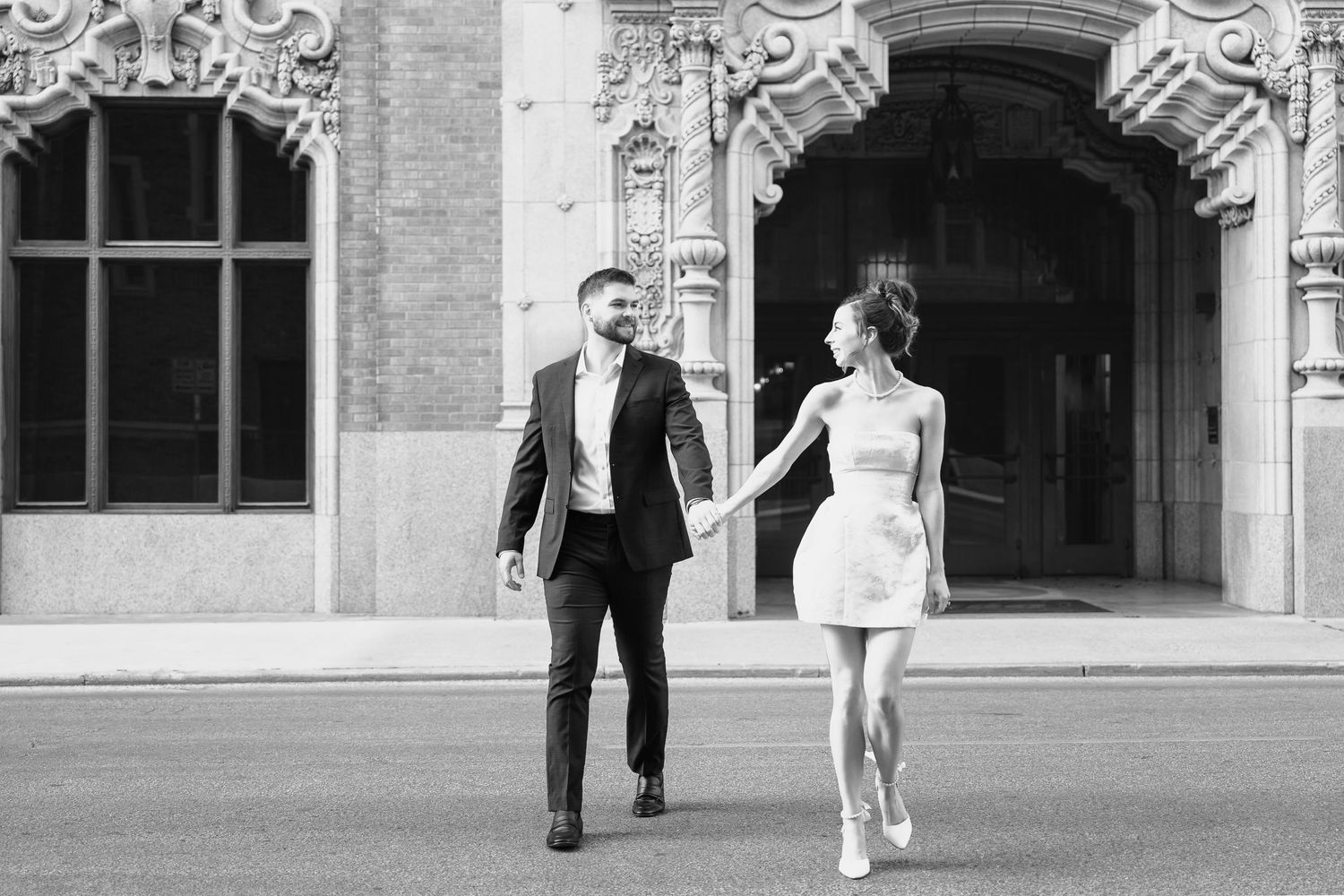 young woman in short white dress holding man in black suits hand and leading him across a downtown San Antonio street