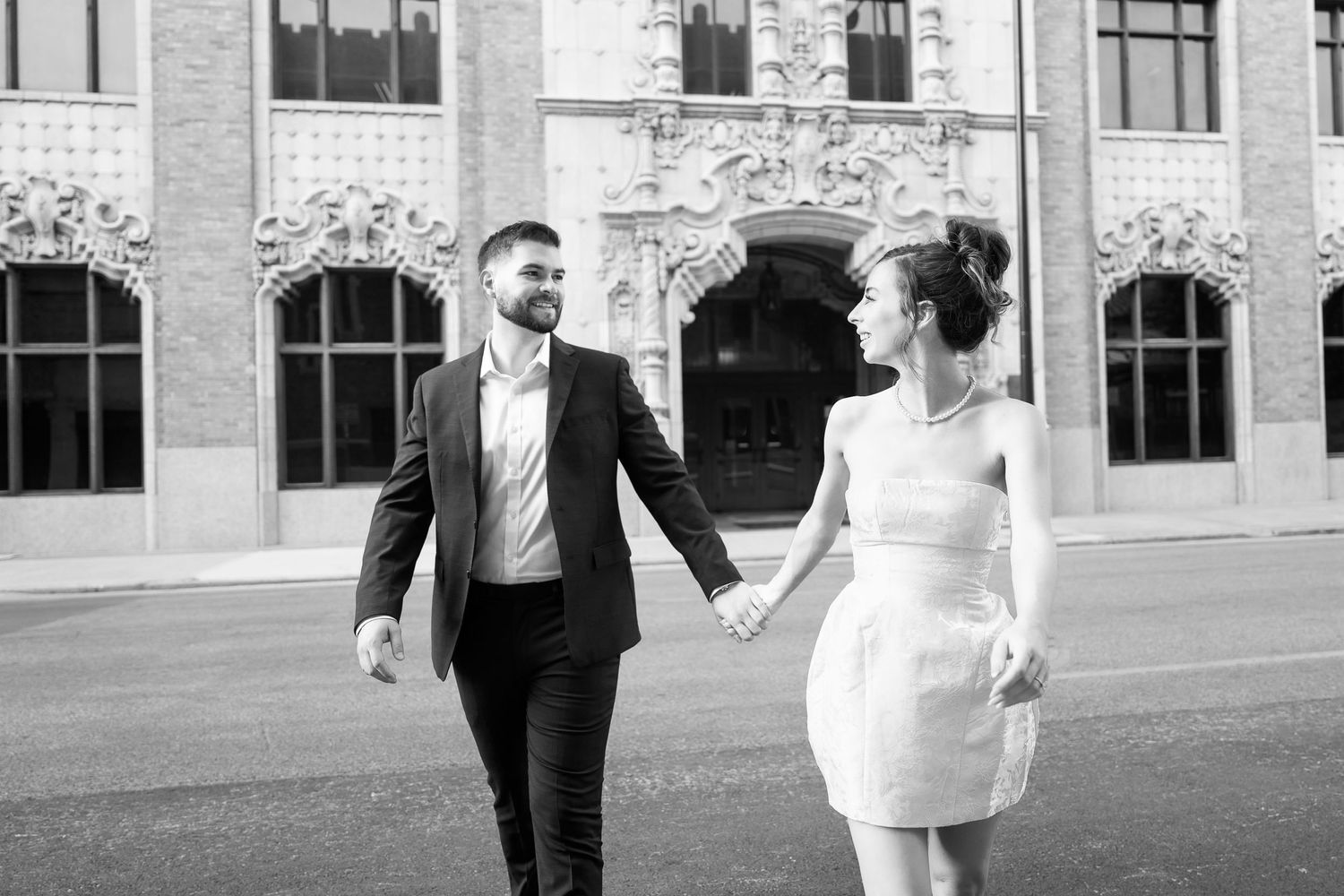 young woman in white dress looking back at her fiancé in a black suit walking across a downtown San Antonio street