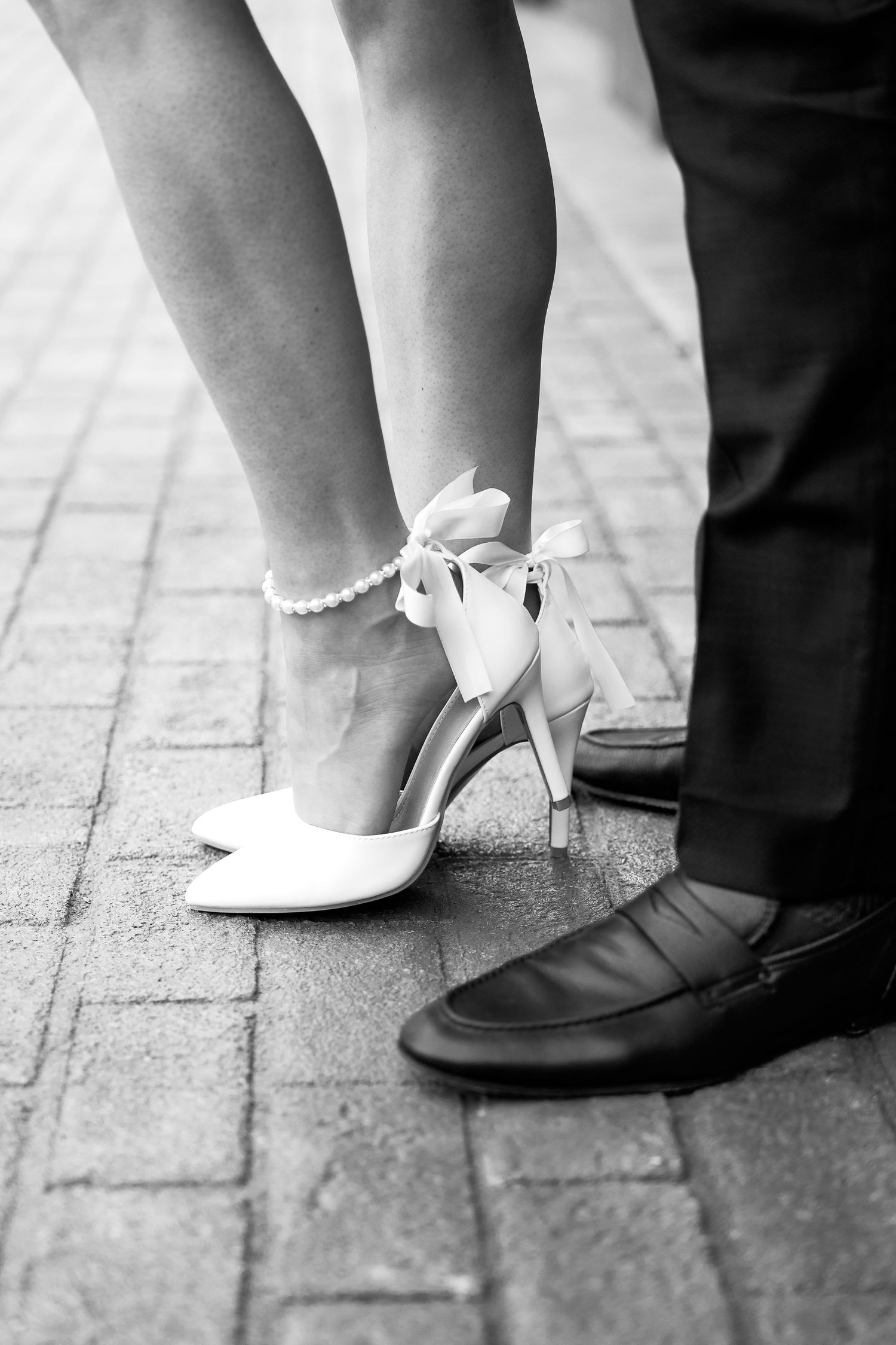 closeup of woman's legs in white high heels with pearls around the ankle and fiancé's black shoes and pants standing
