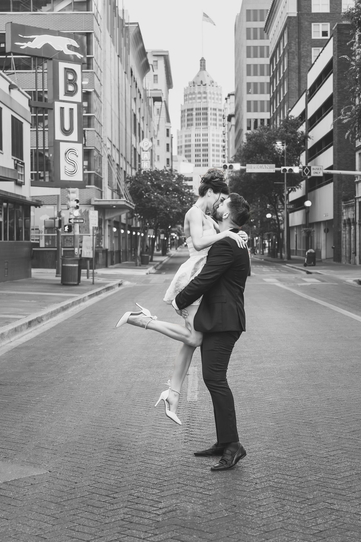 man in black suit picking up fiancé in white dress in middle of downtown San Antonio street with bus sign behind them