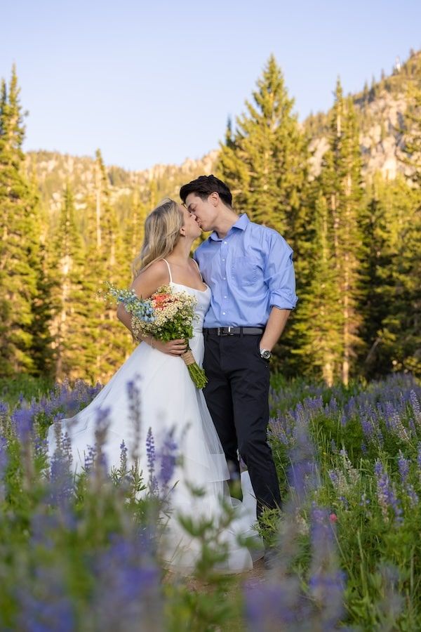 Two people sharing a romantic kiss in a mountain meadow filled with blooming purple wildflowers and evergreen trees.