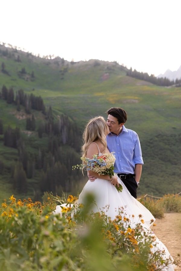 A romantic couple stands among vibrant wildflowers on a mountainside meadow with rolling green hills in the background.