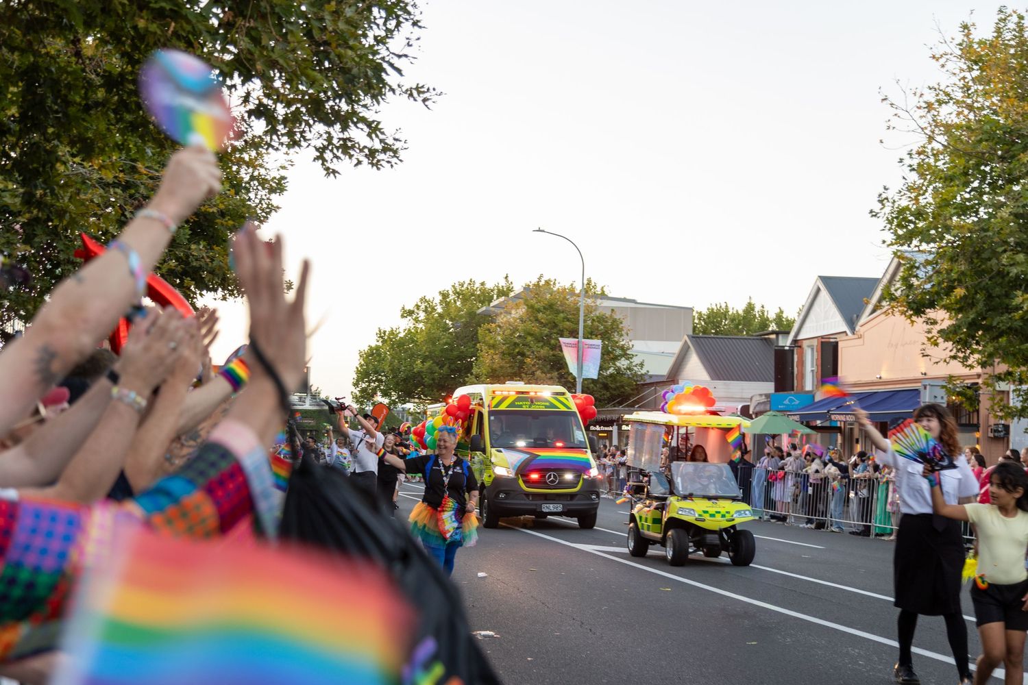 Crowds wave rainbow Pride flags as decorated parade vehicles pass down a tree-lined street at dusk.