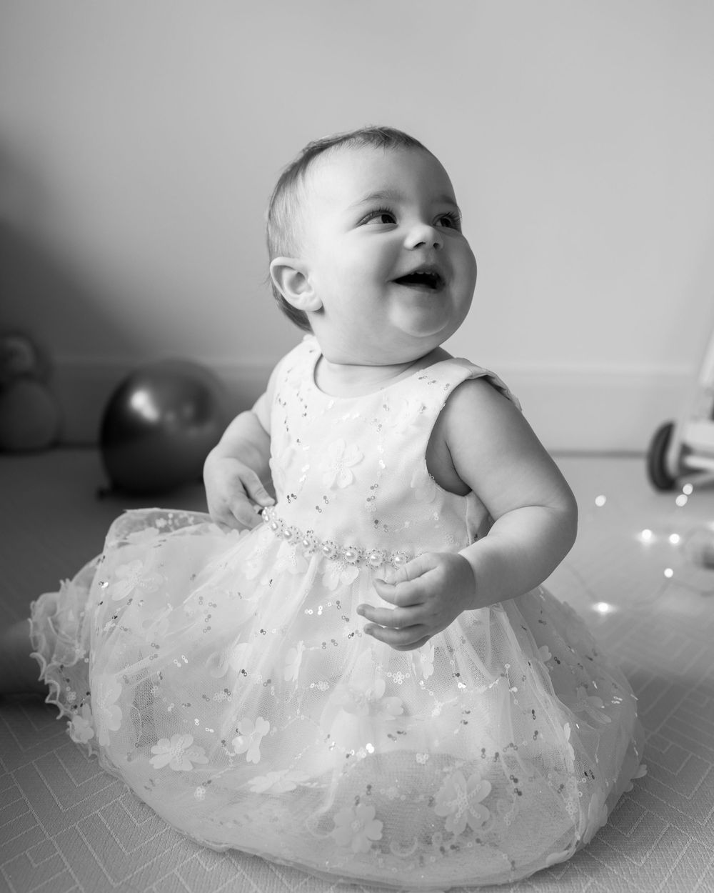 A baby wearing a white tulle dress sits and plays on the floor in a black and white photo.
