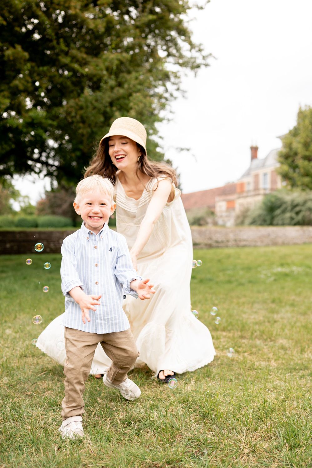 A mother and her son are playing and chasing bubbles together on a grassy lawn with a brick building in the background.