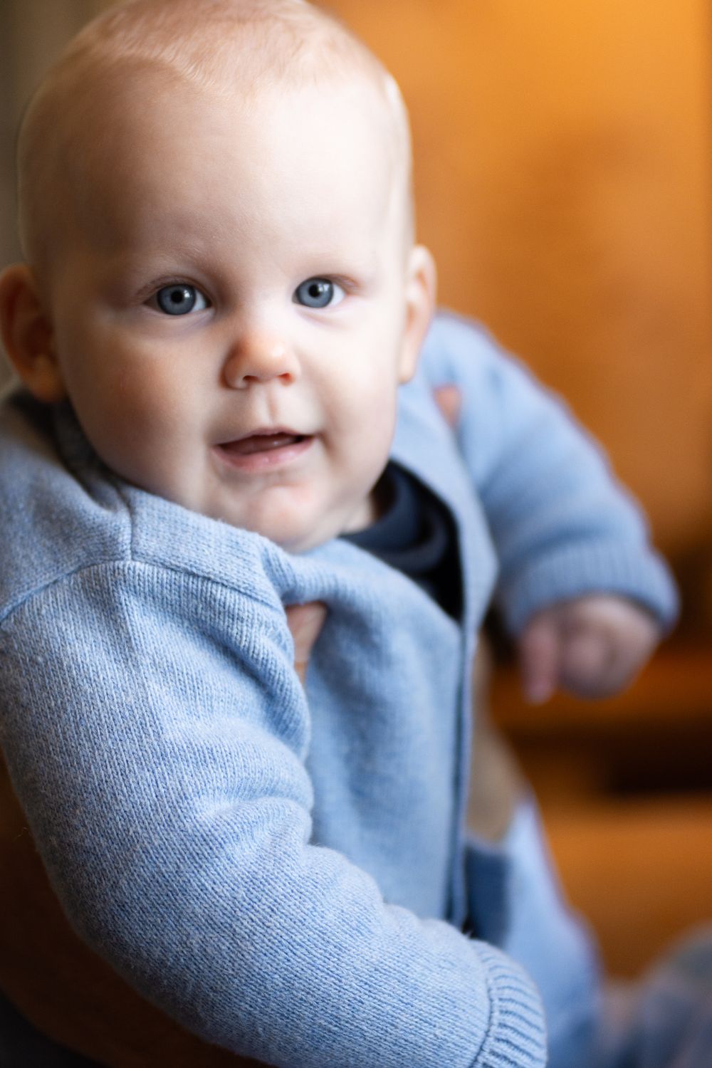 A baby in a light blue sweater smiles while looking to the side with bright blue eyes and soft lighting in the background.