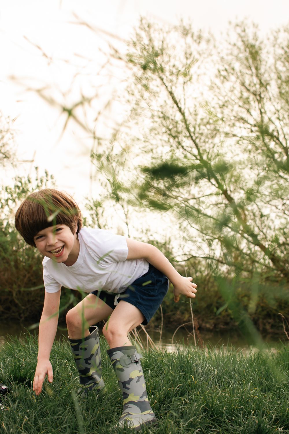 A young child plays and bends down in a grassy field with sunlit trees in the background.