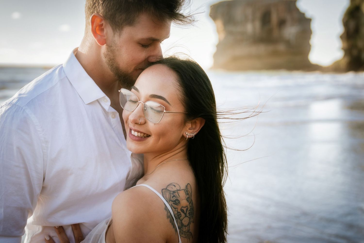 Joyful couple embracing on a beach during sunset with natural Muriwai coastal rocks in background.