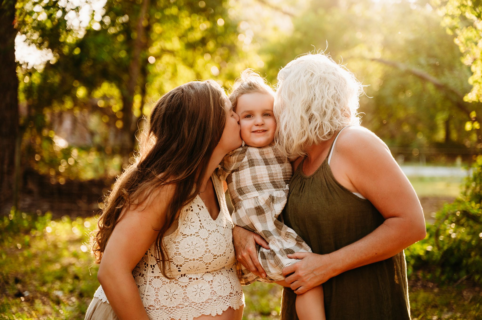 little girl in plaid dress being kissed on the cheek by pregnant mom and grandma