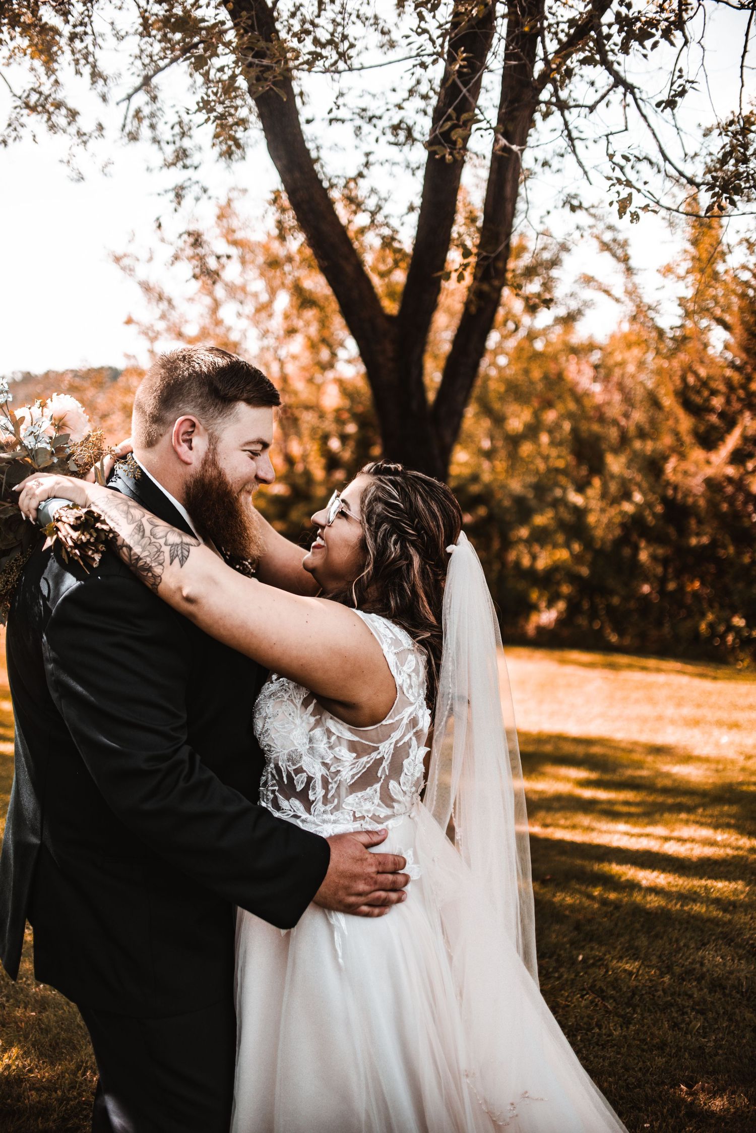 A couple embraces in an autumn setting with golden sunlight filtering through trees during their outdoor wedding.