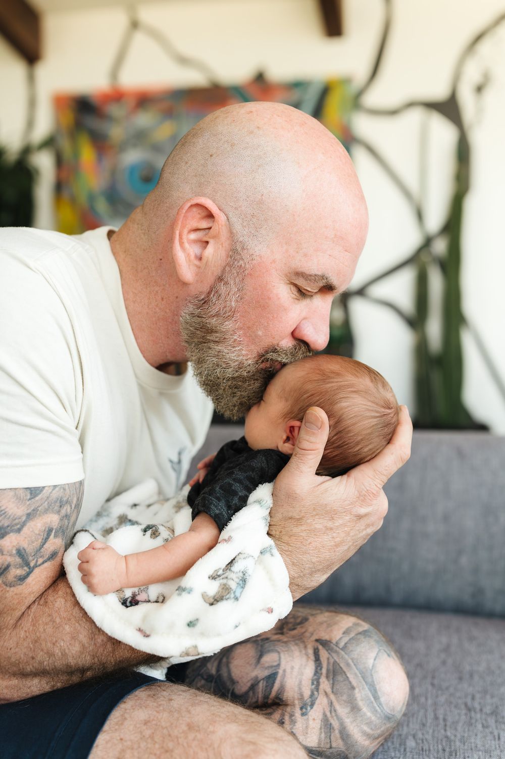 Tattooed bald man with beard tenderly kissing newborn baby while cradling infant in his arms indoors.