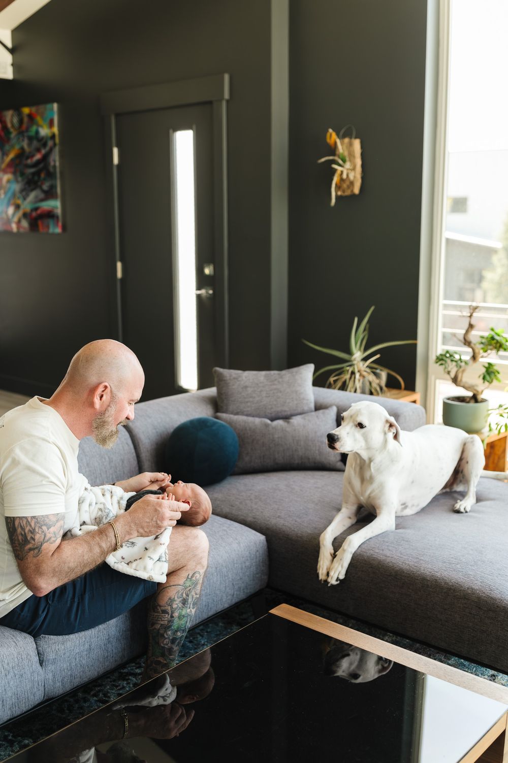 Bald tattooed man holds newborn baby while white dog watches from gray sofa in modern dark-walled living room.