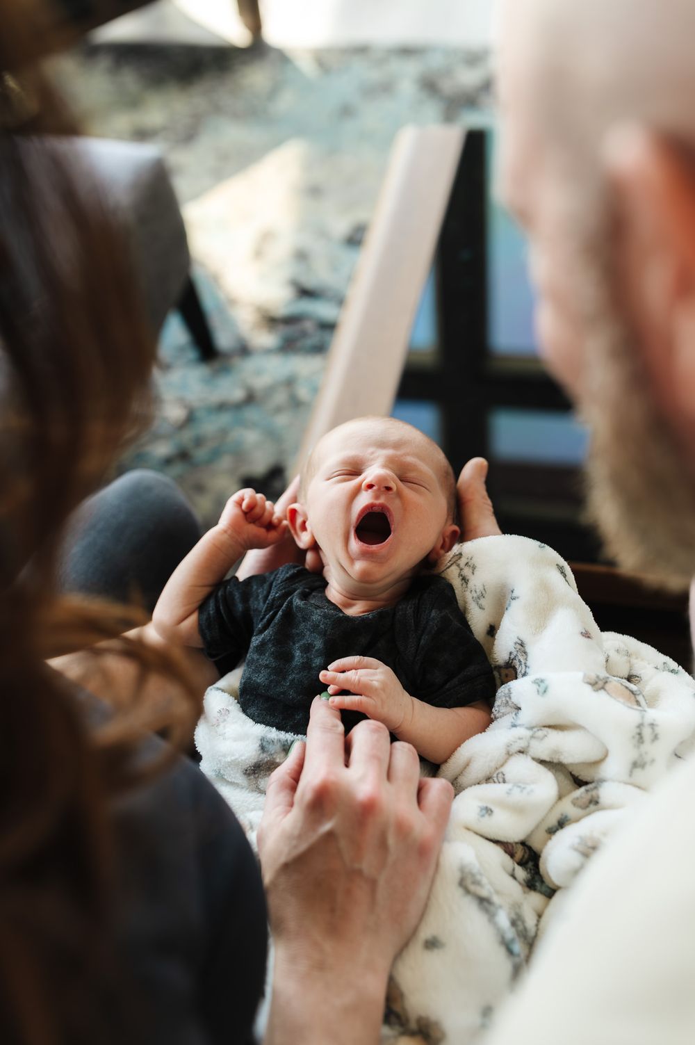 Newborn baby yawning while being held in a blanket by two parents in a cozy home setting.