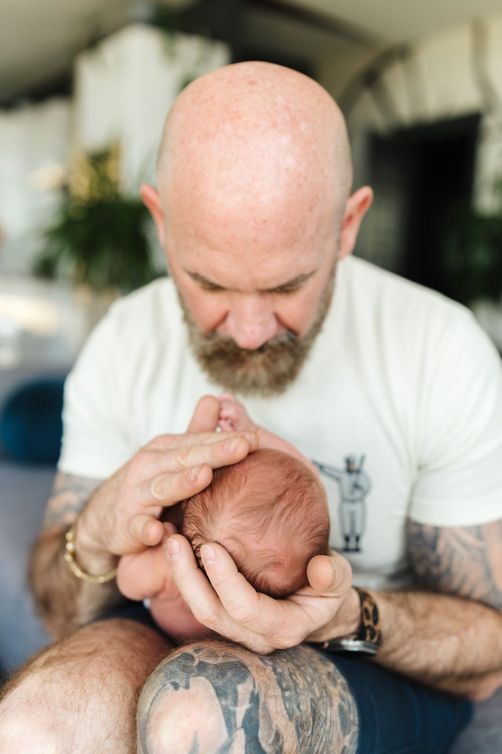 Bald tattooed father with beard gently cradling newborn baby in hands, tender parenting moment.