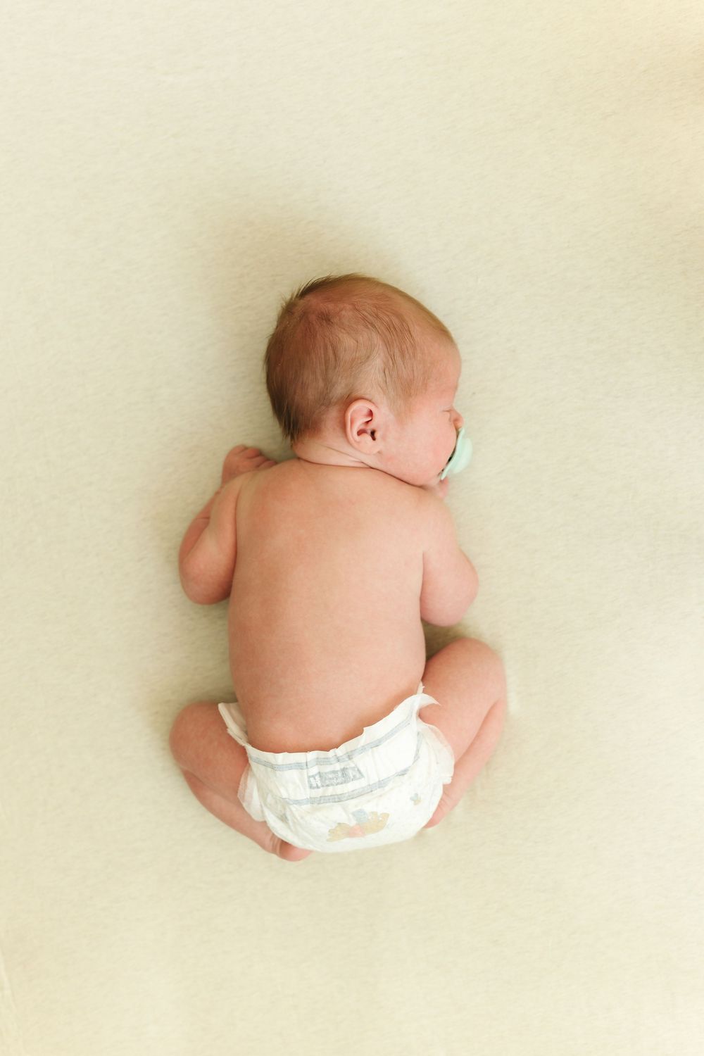 A baby wearing a white diaper lies on a cream-colored background viewed from above.