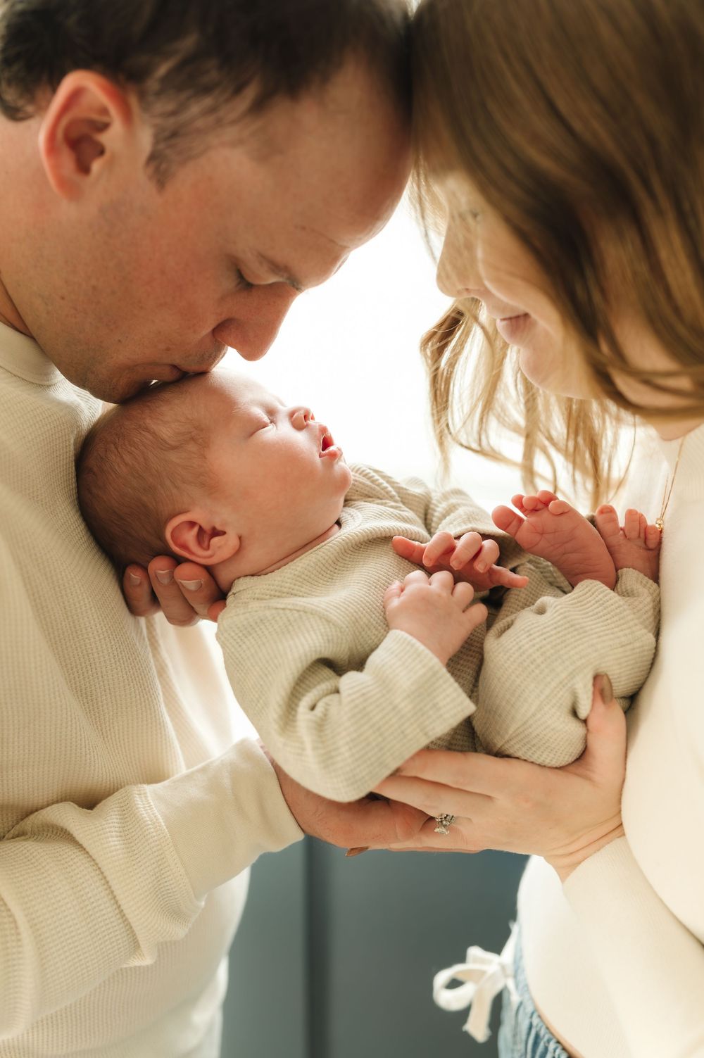 A tender moment as parents in cream sweaters lovingly embrace their newborn baby in a warm, intimate setting.