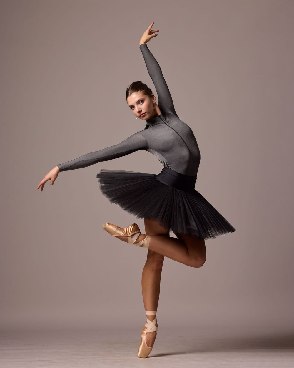 Ballerina in dark leotard and tutu performs graceful arabesque pose on pointe shoes.