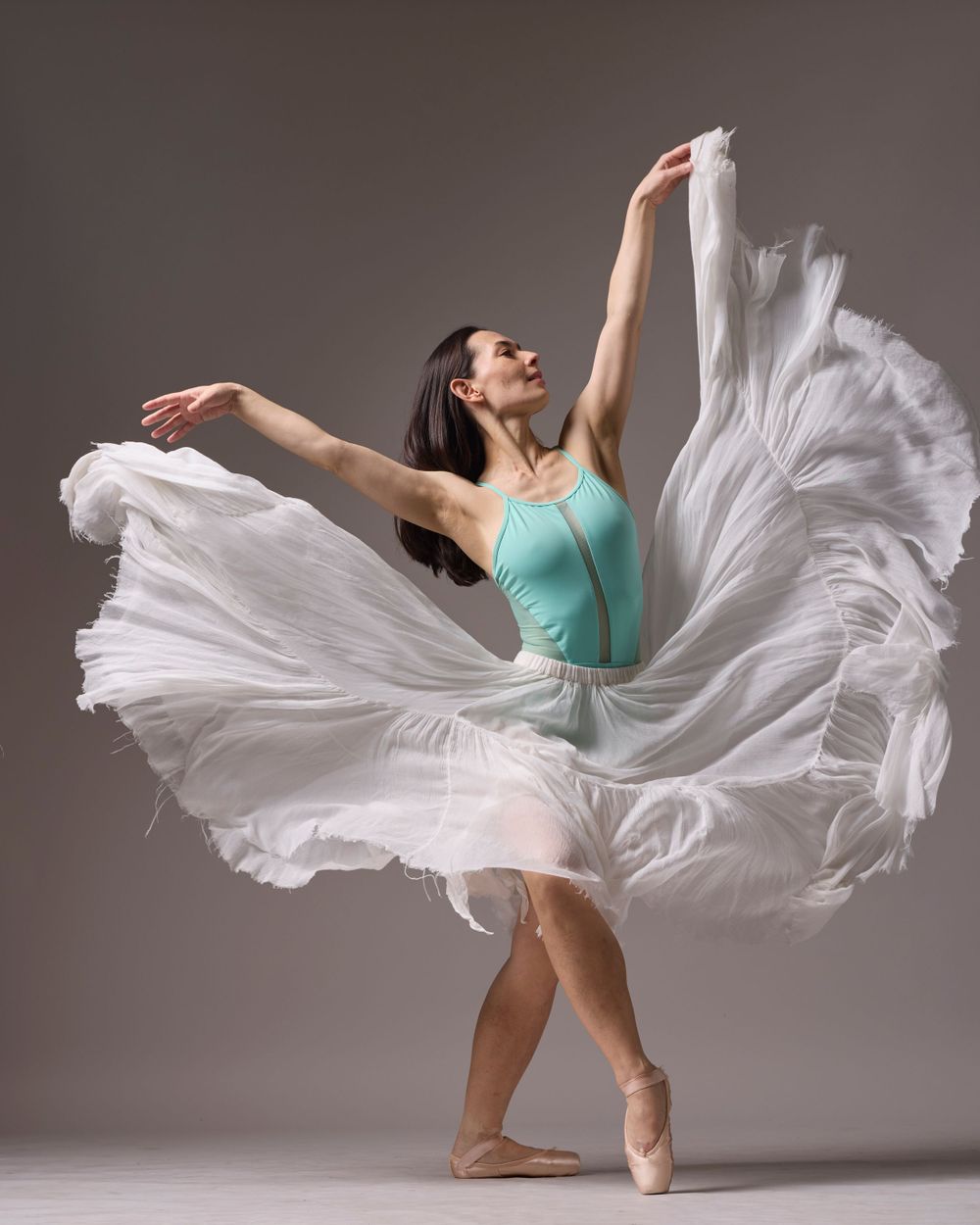 Dancer in turquoise leotard twirling in flowing white dress against studio background.