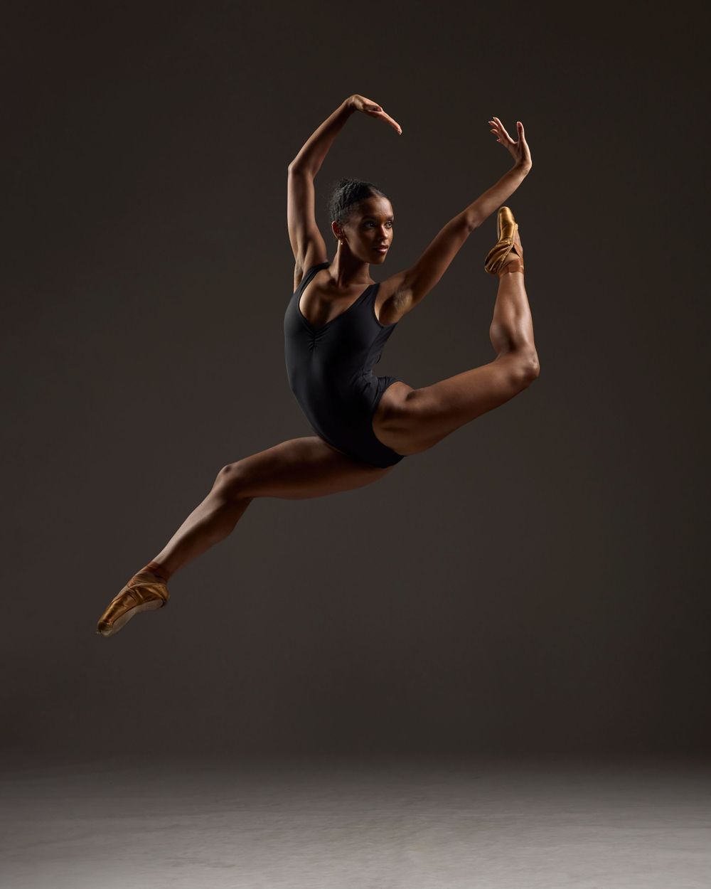 Athletic dancer in black leotard performs dramatic leap with arched back against dark background.