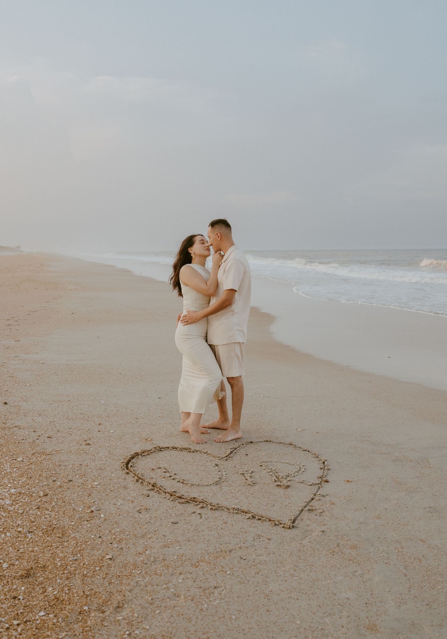 A couple stands inside a heart drawn in the sand on a peaceful beach.