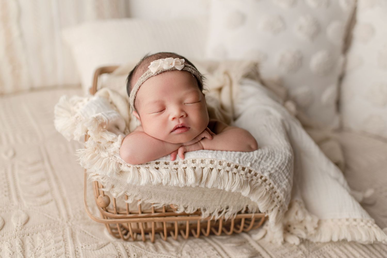 Sleeping newborn baby girl in floral headband posed in wicker basket with cream blanket.