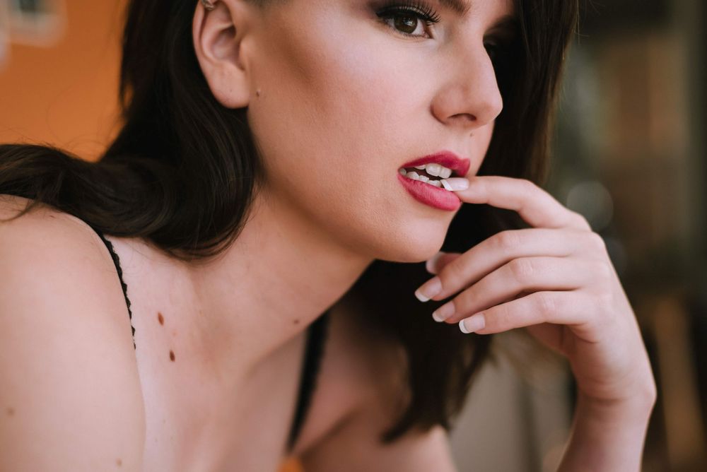 Close-up of a young woman with red lips and dark hair, biting her fingernail in a thoughtful pose.