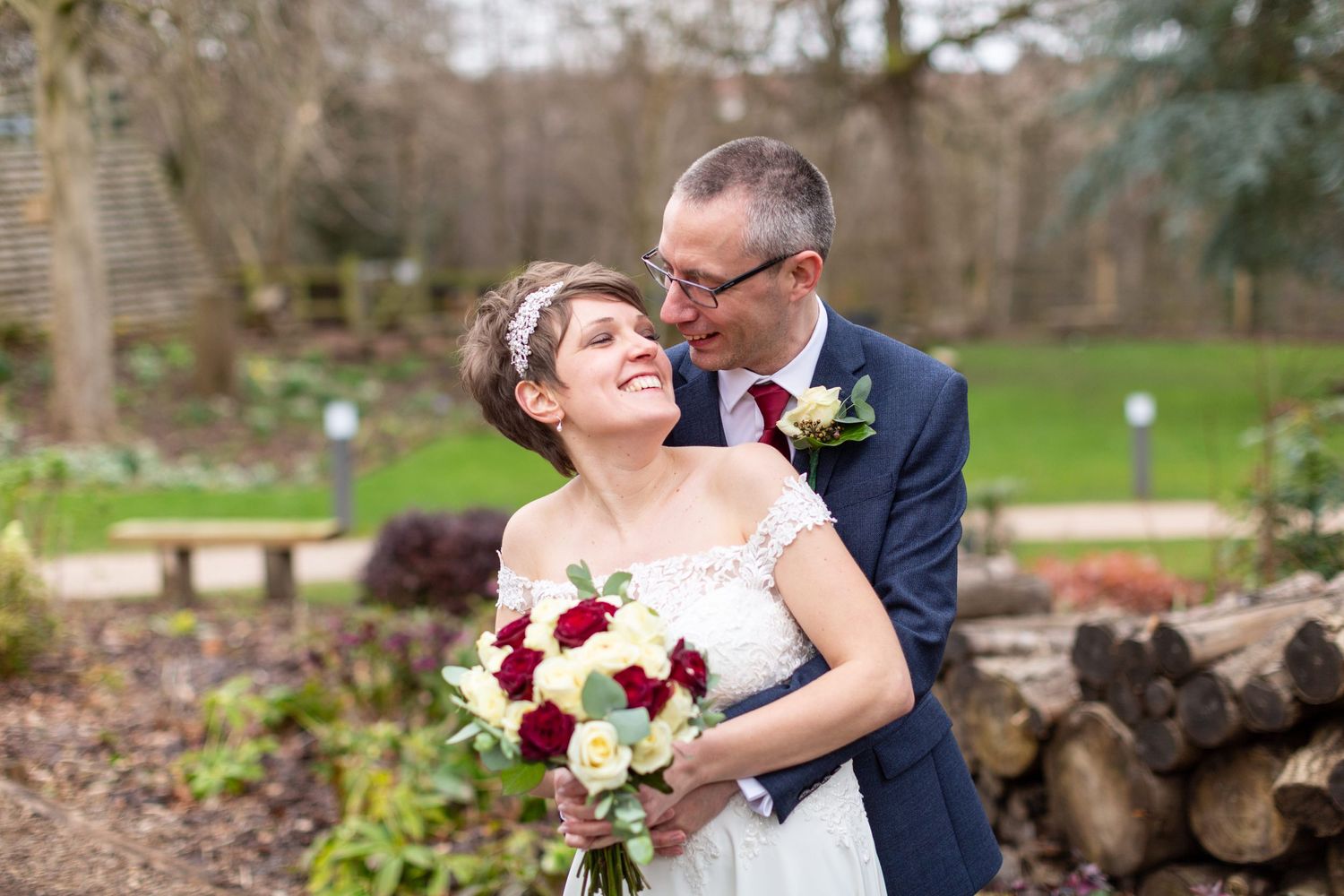 A joyful wedding couple shares a moment in a garden setting with a red and white bouquet.