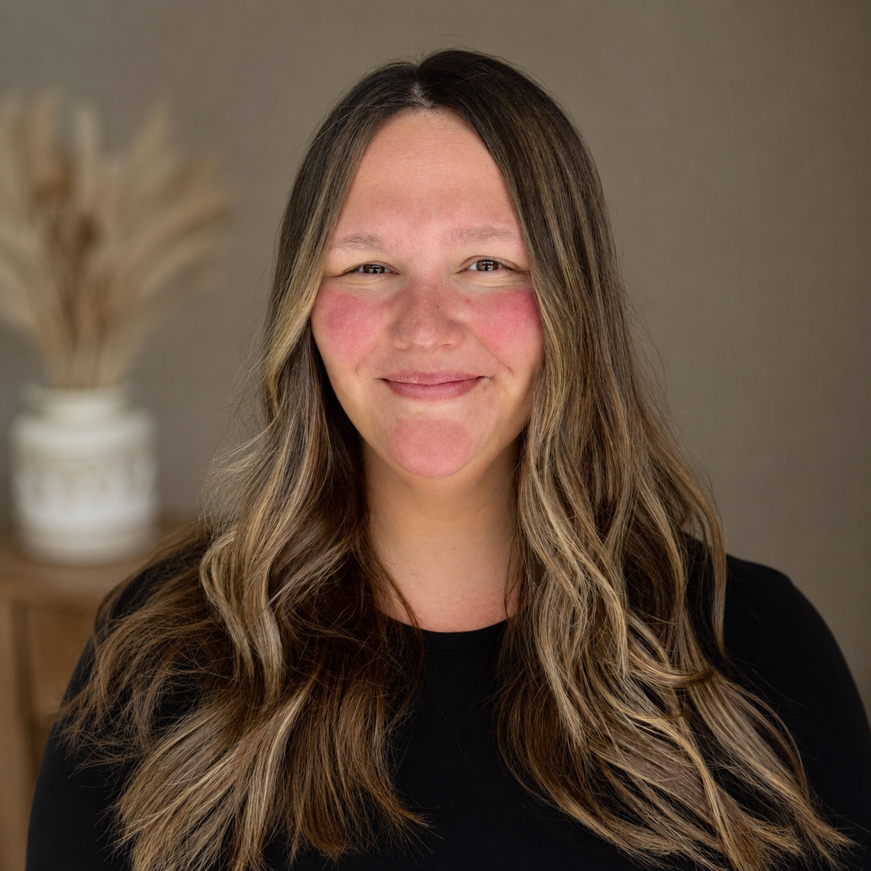 Professional headshot of a smiling woman with long highlighted hair against a warm neutral studio background.
