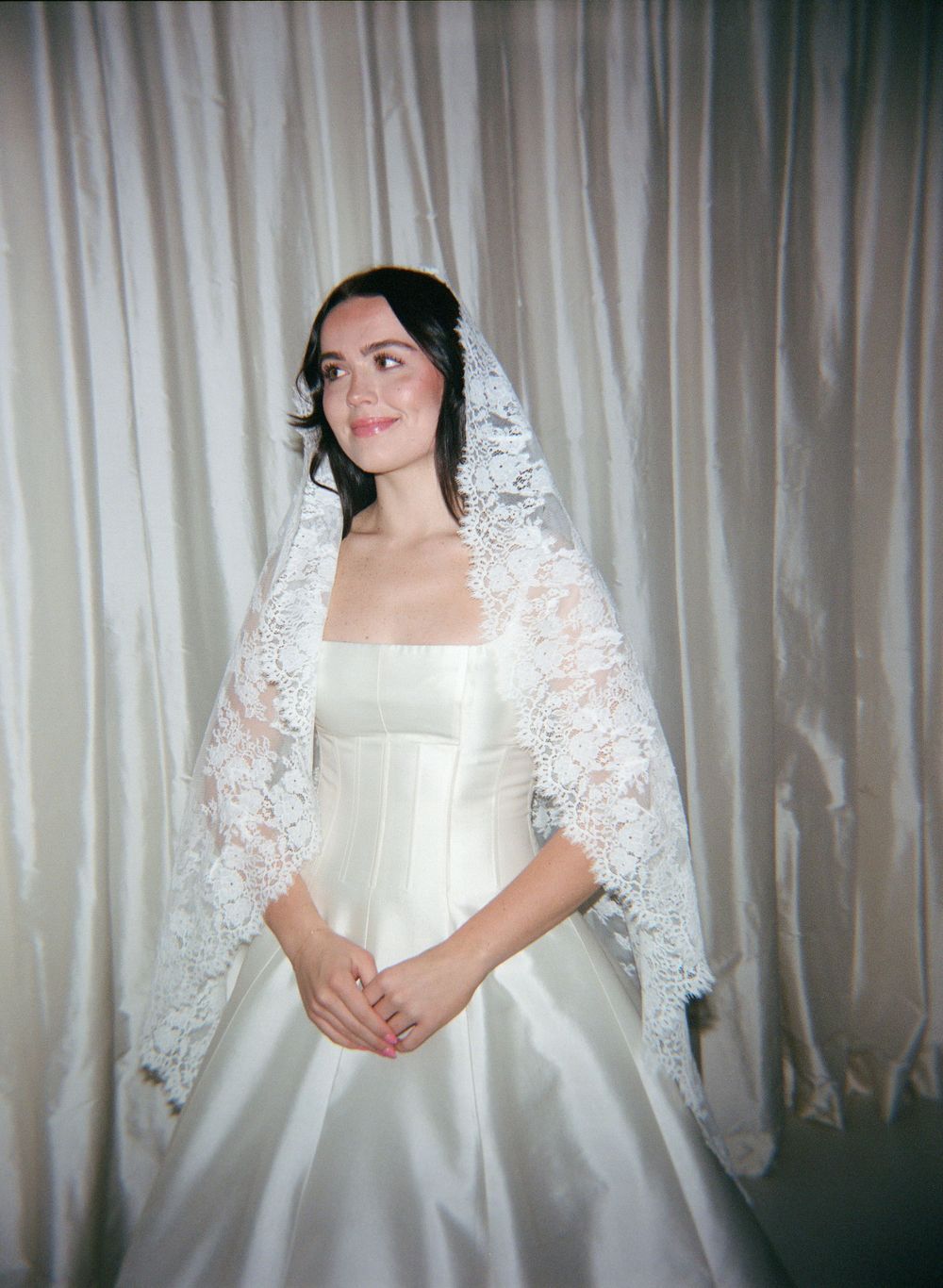 A bride wearing a traditional white wedding dress and delicate lace mantilla veil poses against a light curtain backdrop.