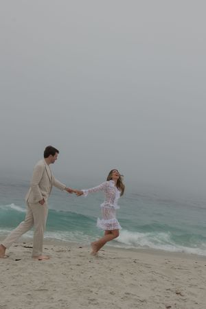 Couple in wedding attire holding hands and dancing barefoot on a misty beach with turquoise waves.