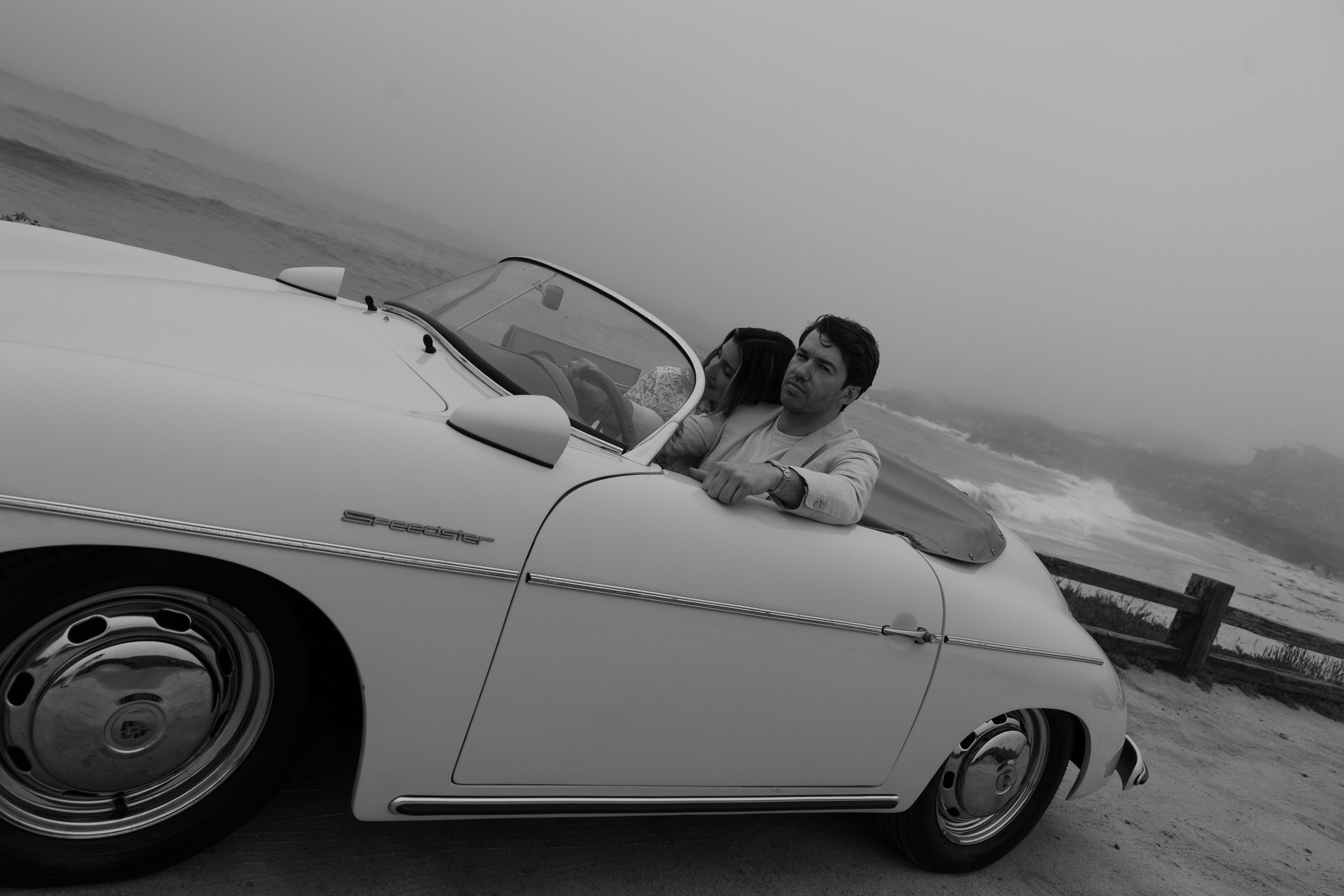 Black and white photo of a couple sharing a romantic moment in a classic white convertible car by the ocean.