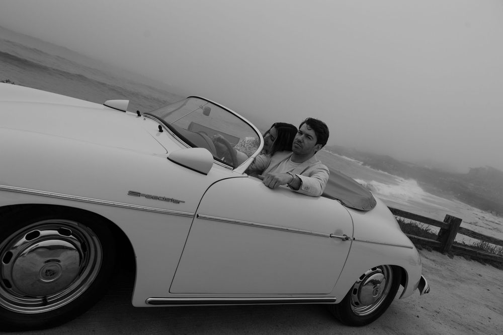 Black and white photo of a couple sharing a romantic moment in a classic white convertible car by the ocean.