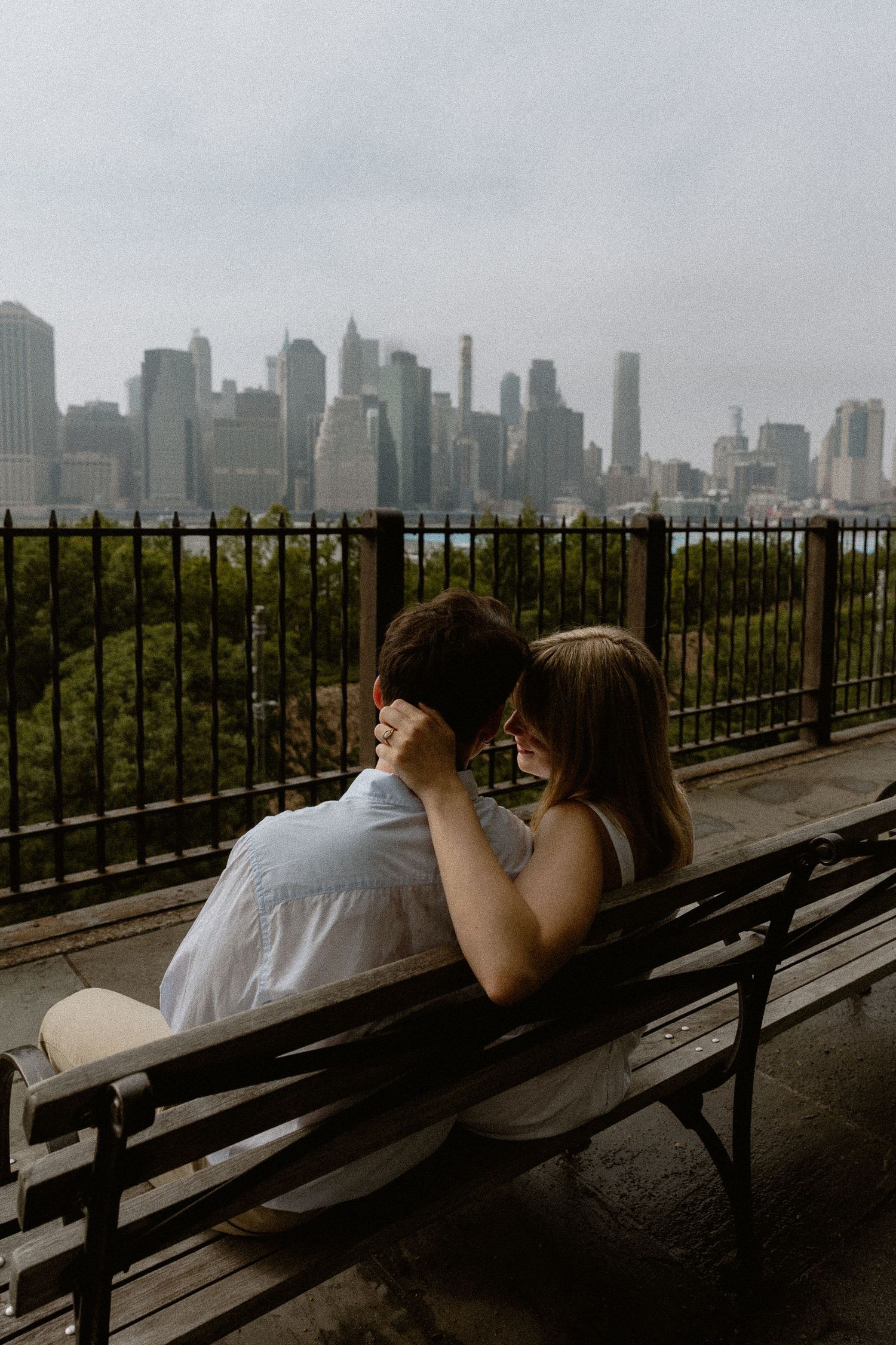 Engaged Couple sitting on a bench sharing a tender moment with NYC skyline backdrop. Brooklyn NY. Miranda Photo & Co
