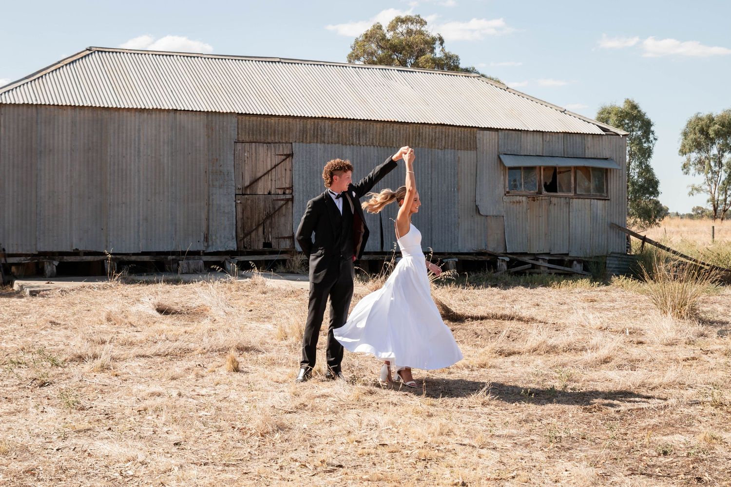 A couple dances together in front of a rustic metal barn building in a dry rural field on their deb day.