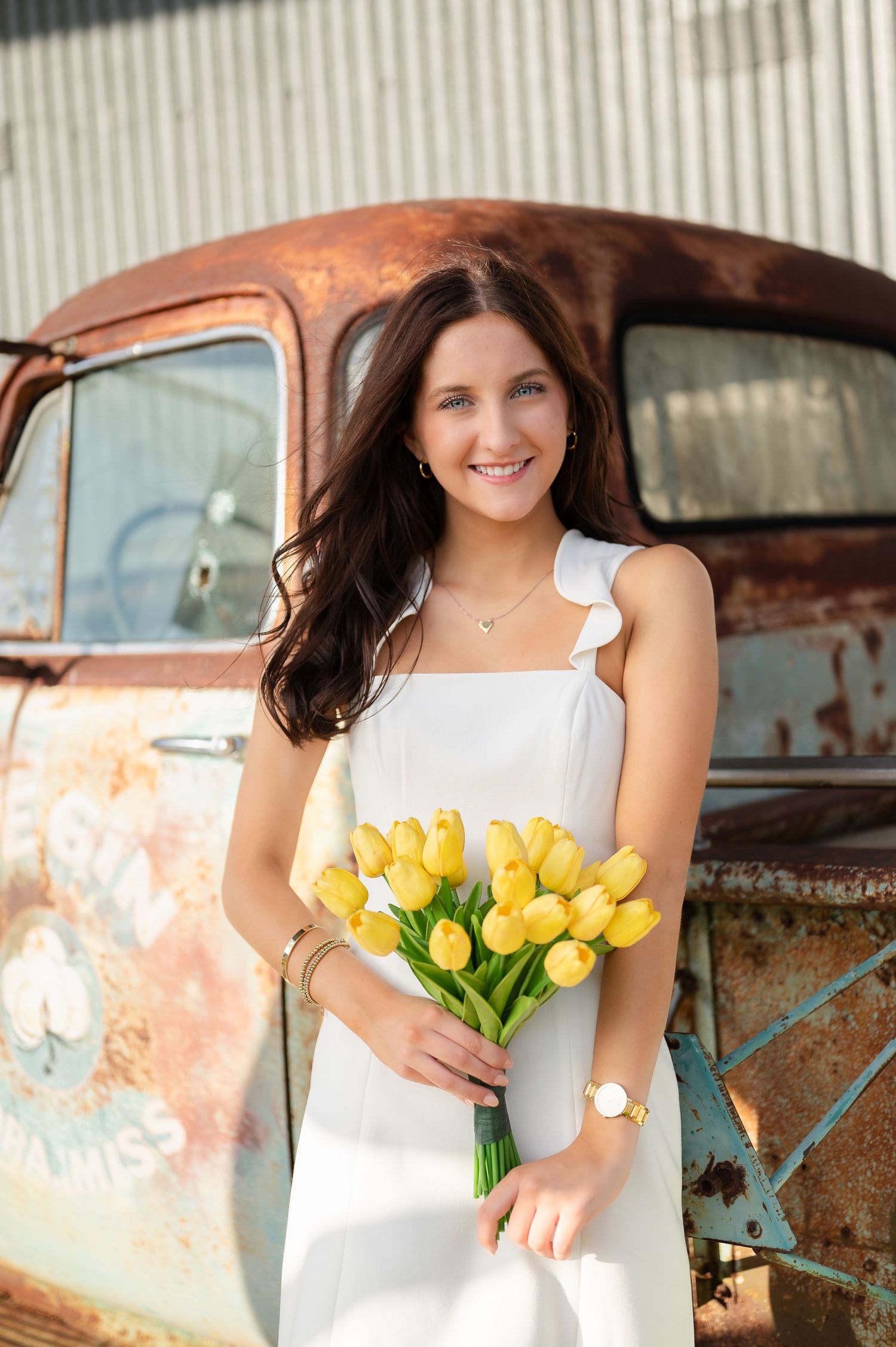 Woman in white dress holding yellow tulips poses by vintage rusty truck.