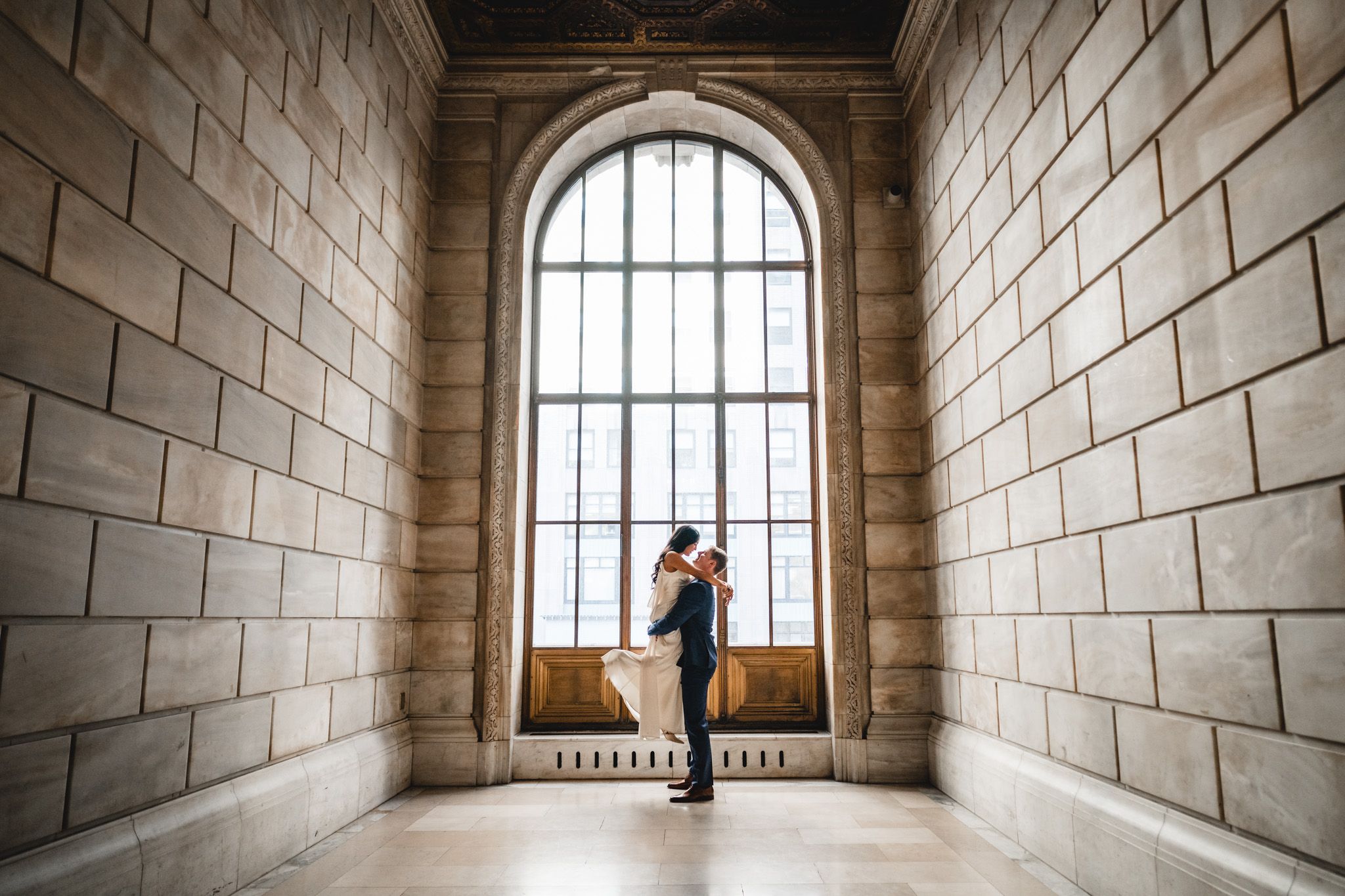a romantic backlit portrait at New york public library engagement session the groom lifting the bride in the large window corridor