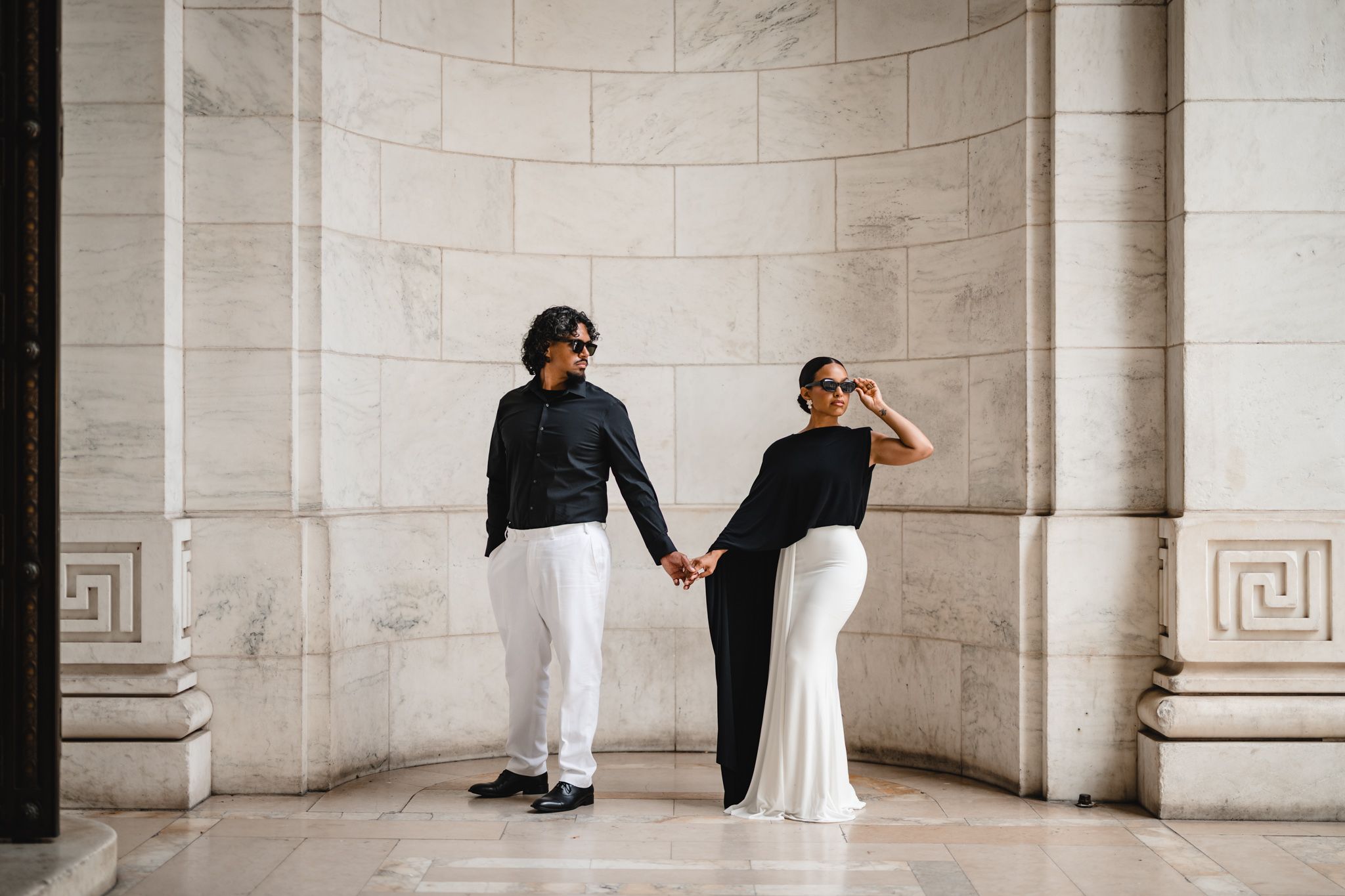 Elegant couple in black and white outfits holding hands by marble columns in a grand architectural setting.
