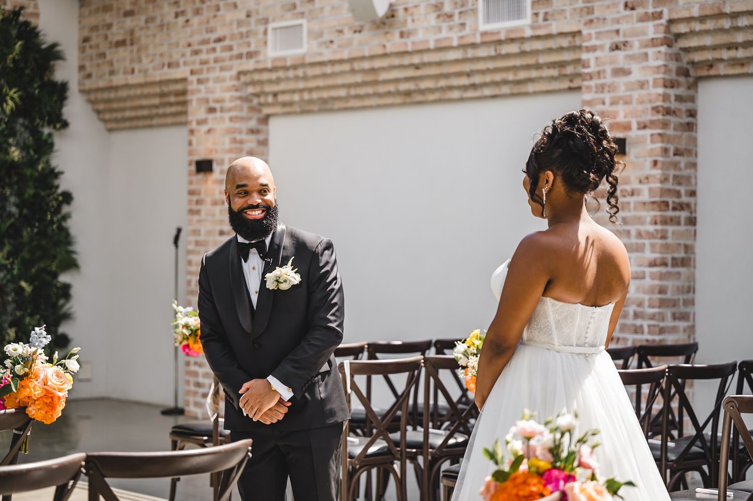 A joyful wedding moment captured outdoors with brick walls and cross-back chairs in the background during first look perona farms refinery