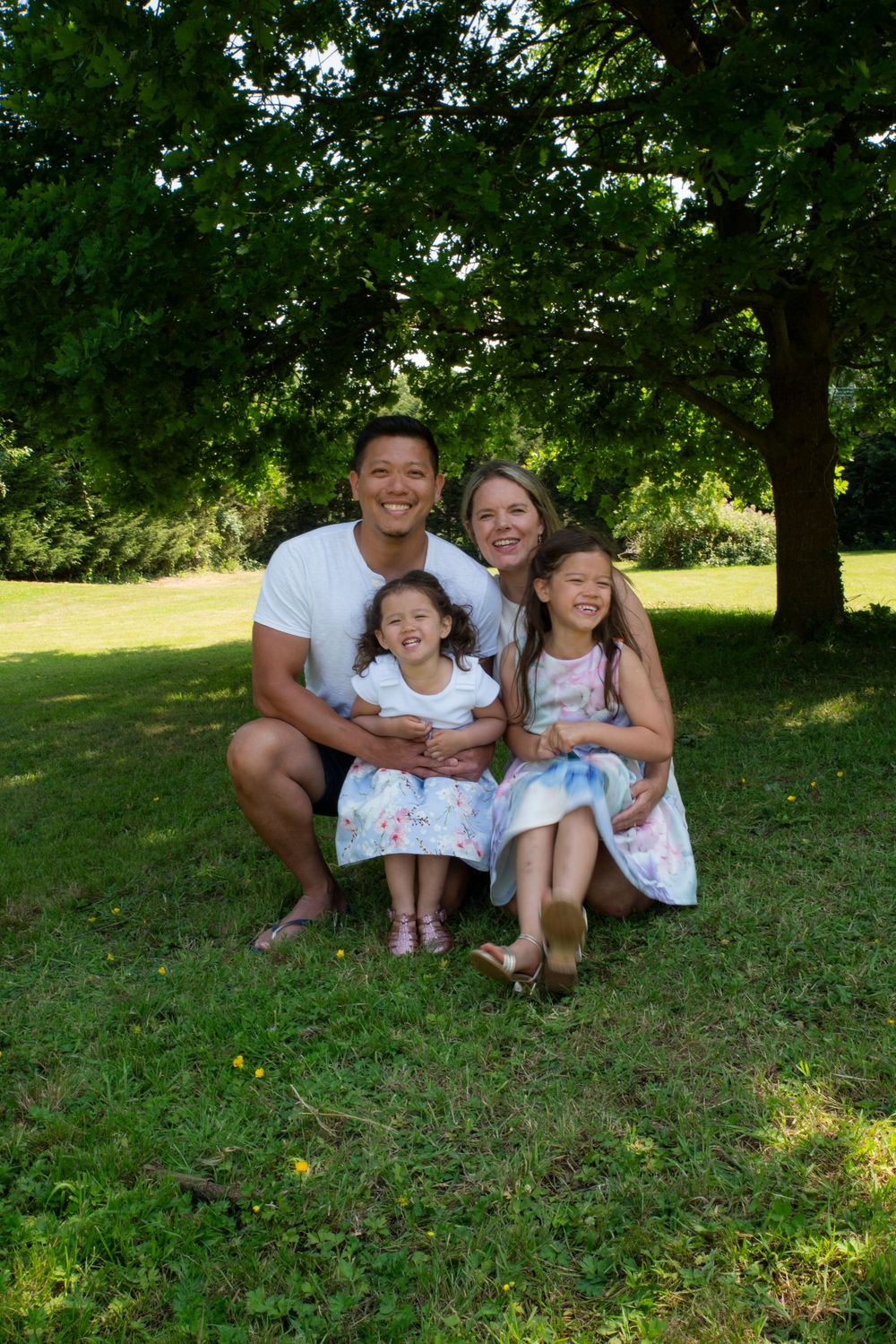 Family sits together on grassy hillside under shady trees on a sunny summer day.