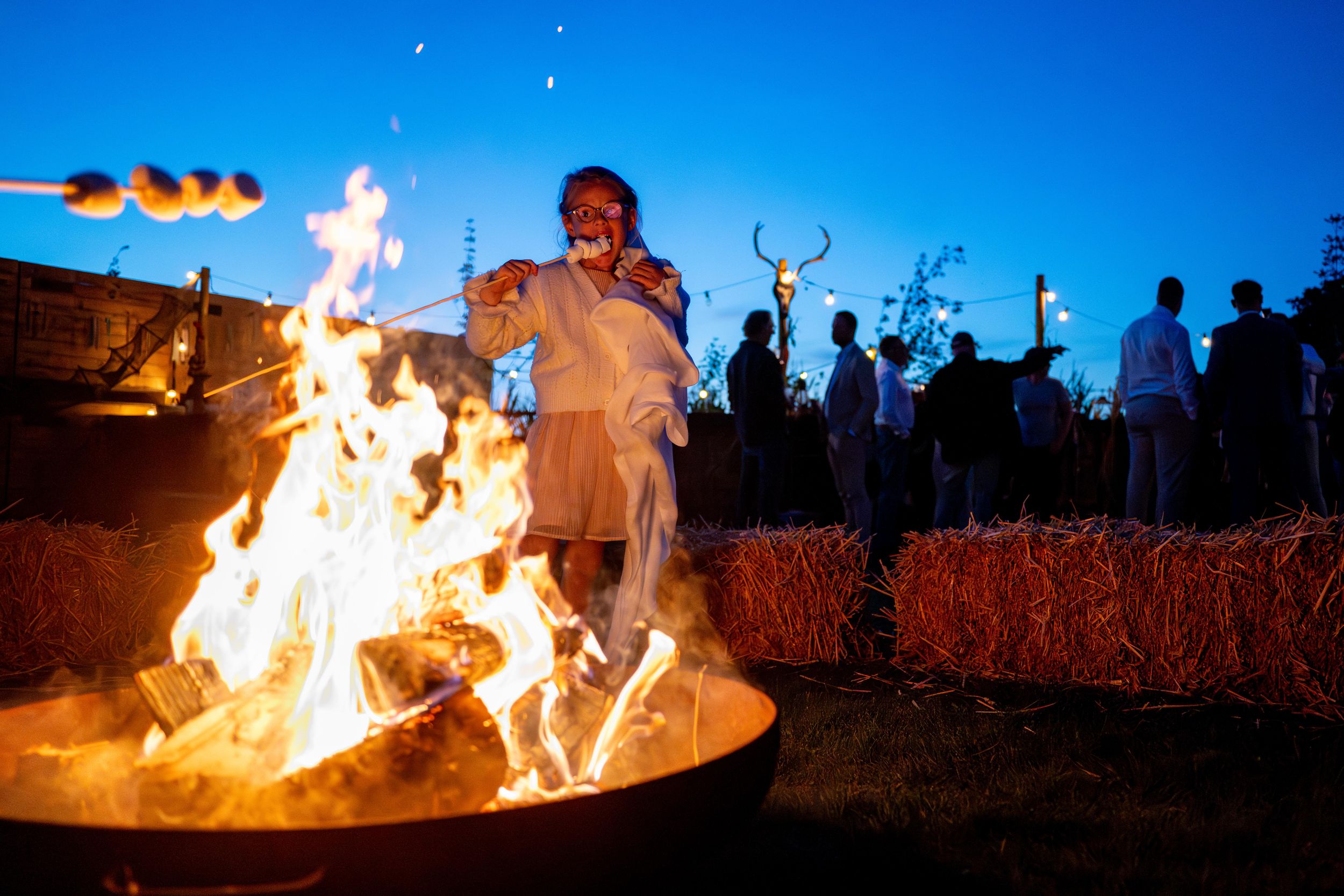People gather around a large fire pit at dusk, with hay bales and deer antler decorations in the background.
