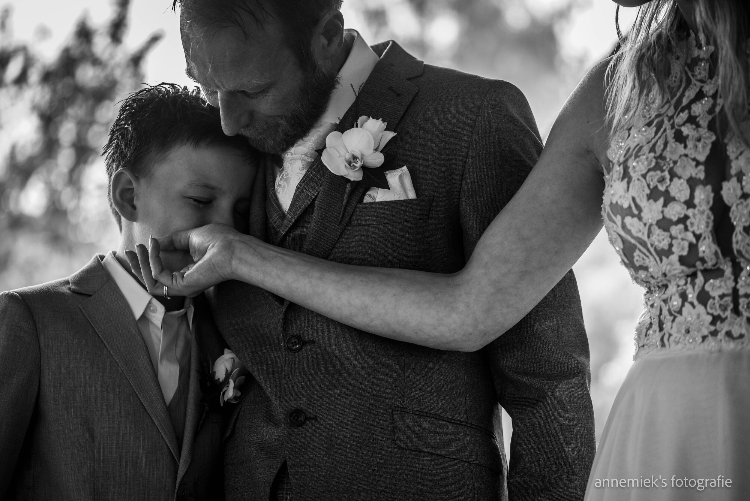 Black and white wedding photo of groom embracing young boy while bride tenderly places hand on their shoulders.