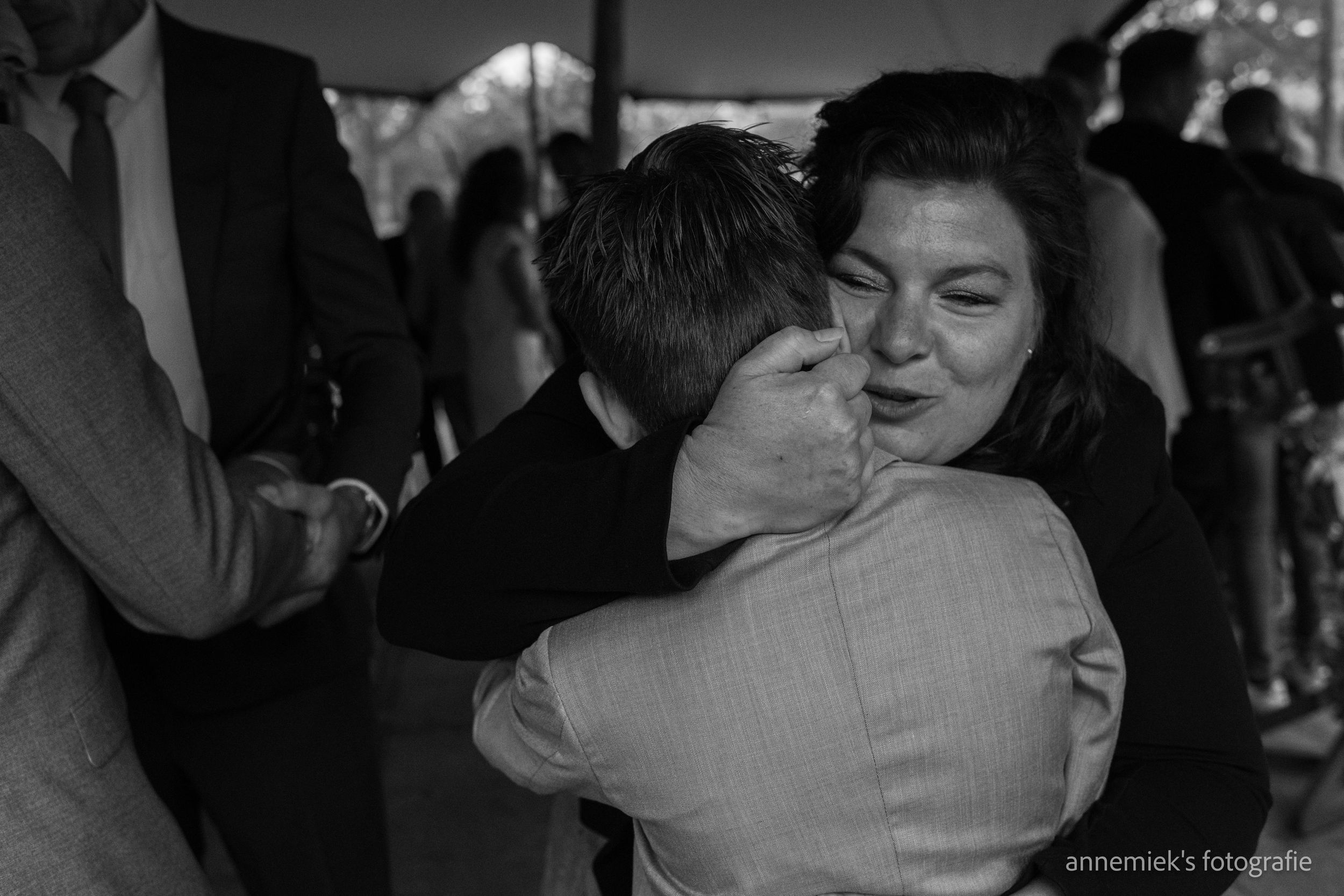 Black and white photo of two people sharing a warm embrace and kiss at a social gathering or wedding reception.