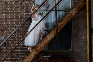 Bride in white tulle dress poses gracefully on rustic metal fire escape stairs against a brick building wall.