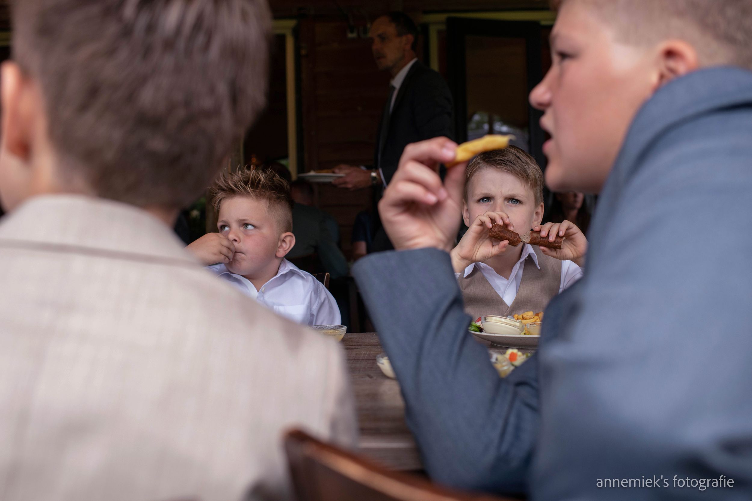 Children eating snacks at a formal event, with boys in suits enjoying food at a wooden table.