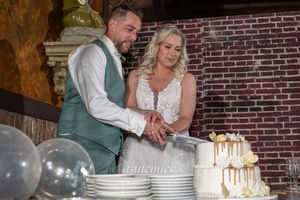 Wedding couple cuts their elegant white and gold cake at reception with brick wall backdrop.