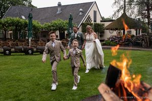 Two boys in suits run toward a bonfire as a bride and groom follow on a green lawn outside a wedding venue.