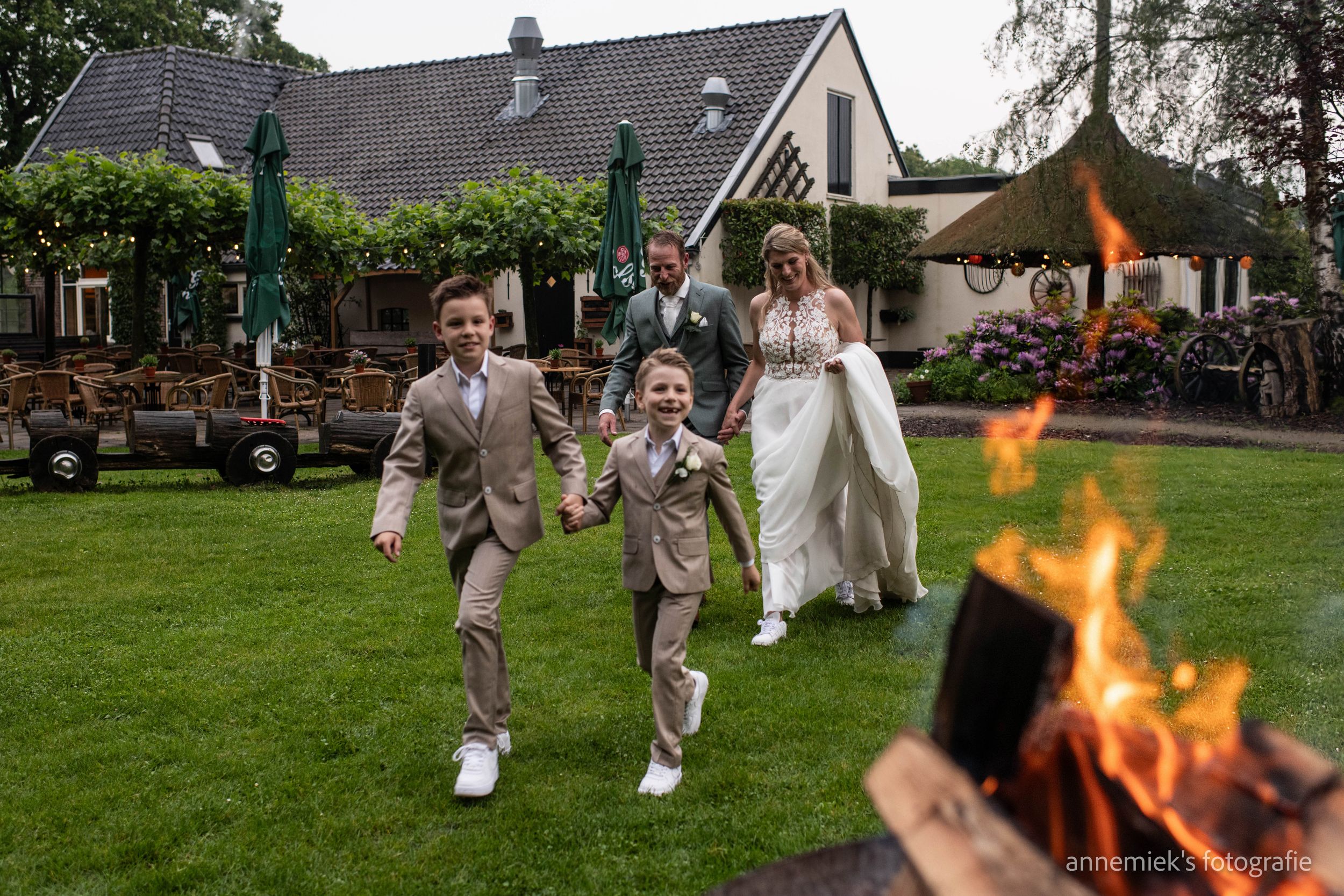 Two boys in suits run toward a bonfire as a bride and groom follow on a green lawn outside a wedding venue.